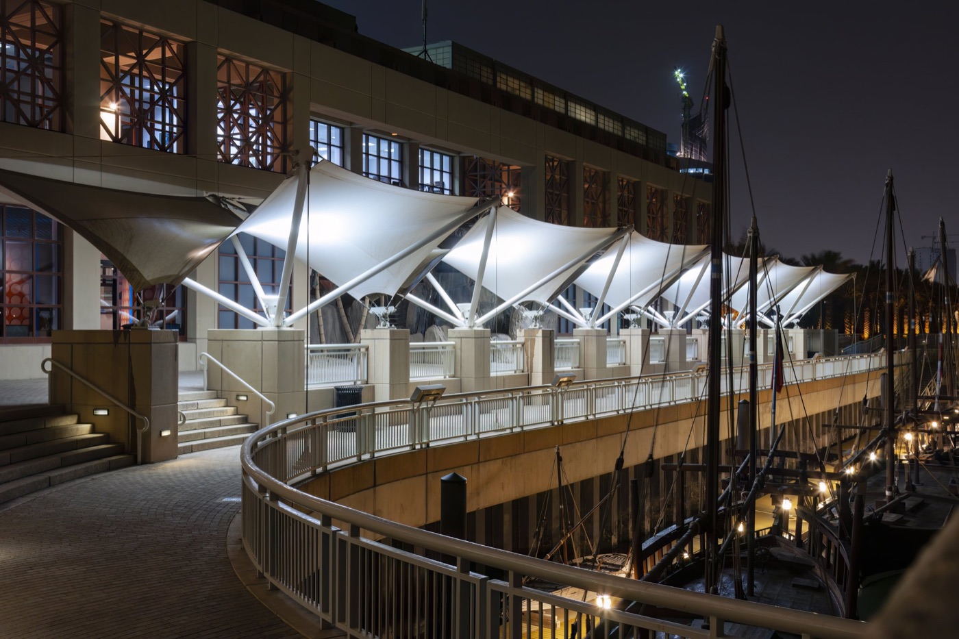 The Scientific Center of Kuwait at night, with white tensile canopies above a curved walkway beside moored wooden boats