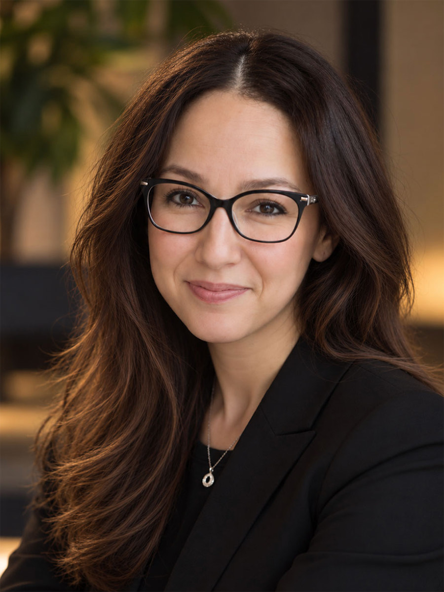 Portrait of Sarah Al-Karmi with long dark hair and glasses, wearing a black blazer
