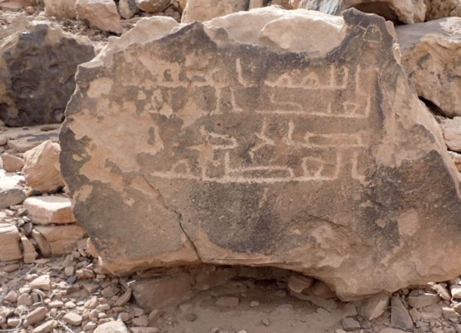 A weathered stone block carved with a Kufic-style Arabic inscription rests in a desert clearing, documented during the Hajj and Trade Routes archaeological survey in NEOM, Saudi Arabia.